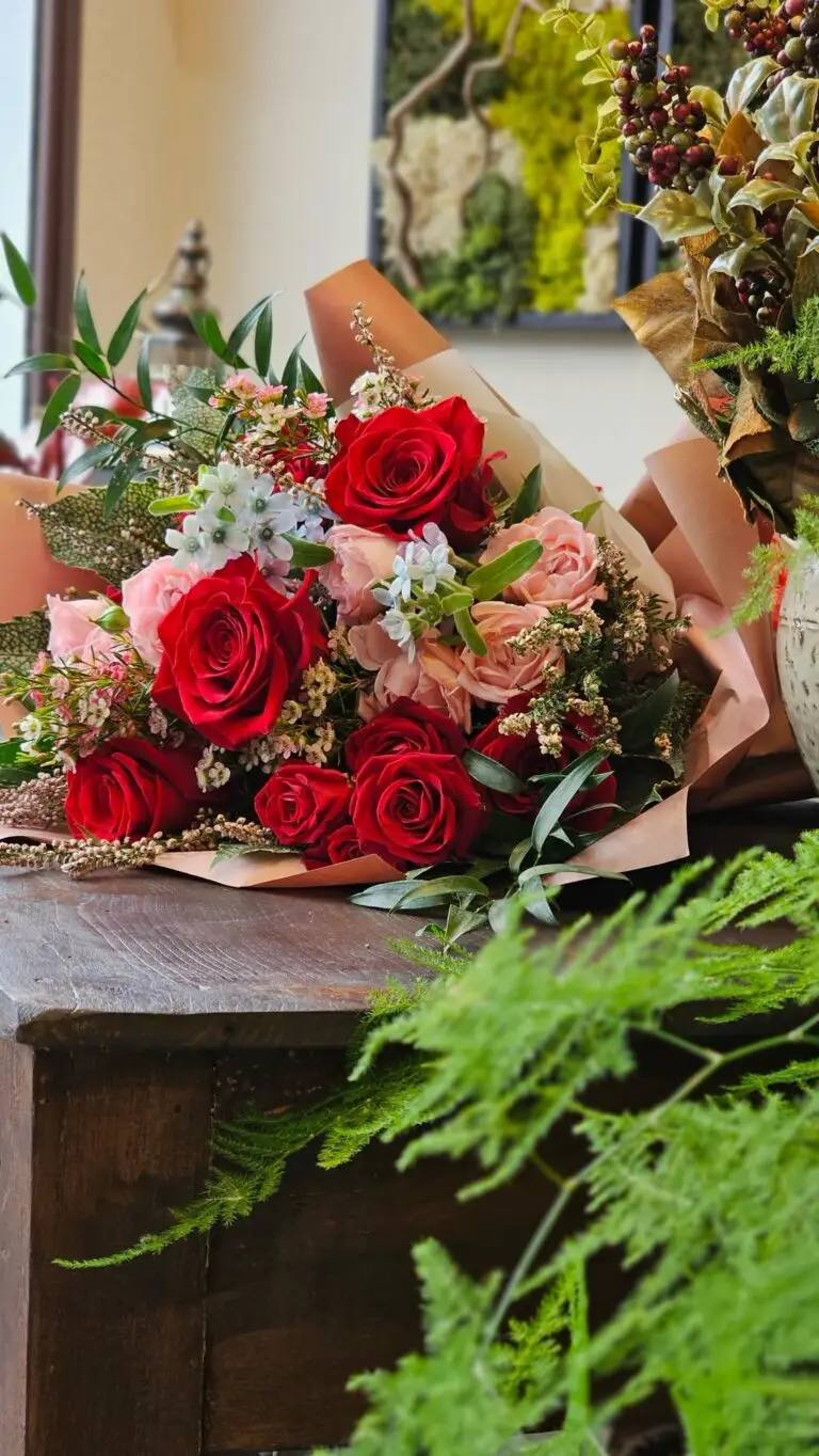 Bouquet di rose rosse e rosa con fiorellini bianchi, incartato e appoggiato su un mobile di legno scuro