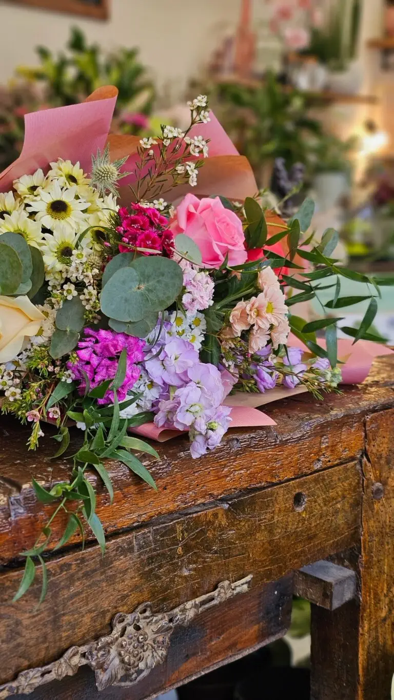 Bouquet di margherite bianche, rose fucsia e fiori lilla con eucalipto, appoggiato su un tavolo di legno antico