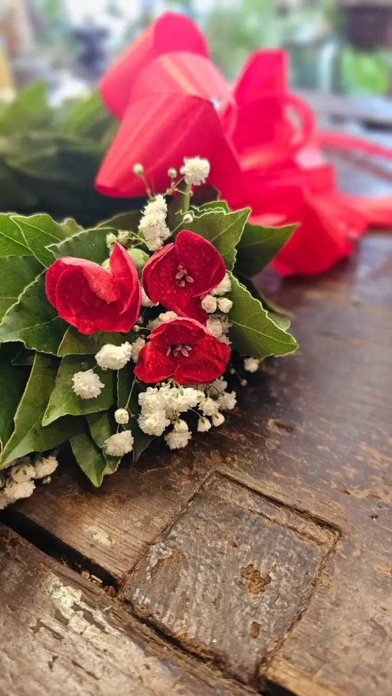 Corona di laurea in foglie di alloro con anthurium rossi, fiorellini bianchi e fiocco di raso rosso, su un tavolo di legno