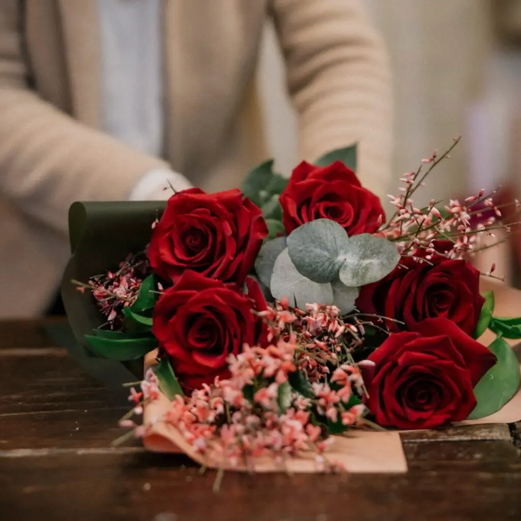Mani che sistemano un bouquet di rose rosse con eucalipto e fiorellini rosa su un banco di legno